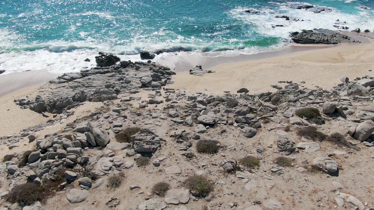 A second view of Buzzards Beach shows white surf spreading across the upper sand while rock clusters fill the lower foreground and no visible people appear.