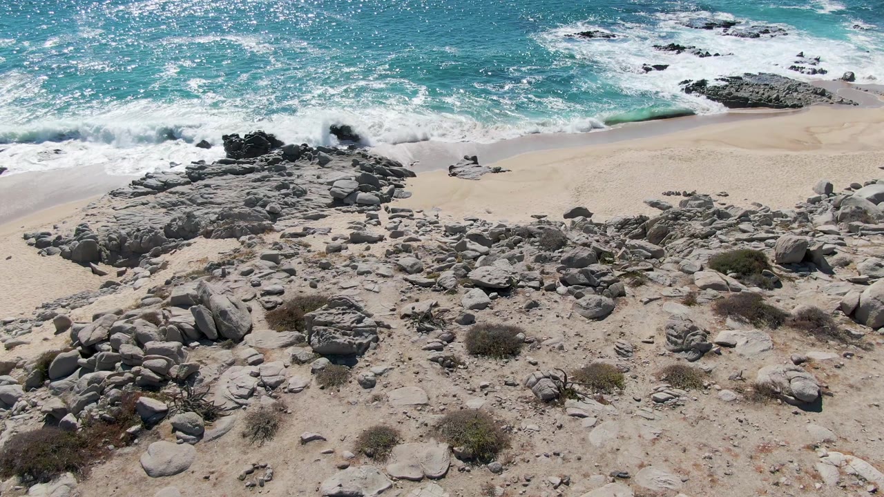 The Baja California Sur shoreline remains empty as blue-green water foams around offshore rocks and pale sand curves behind the rocky foreground.