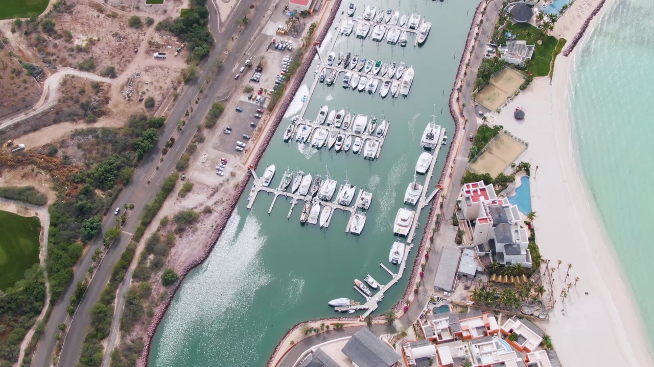 Overhead La Paz marina frame with a small white boat near the dock area, rows of moored yachts, cars on the roadside, a curved beach, turquoise coastal water, and no visible people.