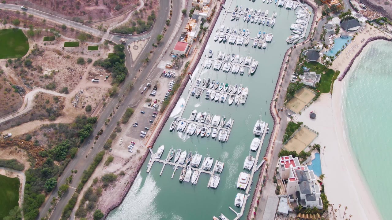 Overhead view in La Paz, Baja California Sur showing a coastal road, dry hillside, marina boats, pale turquoise water, sandy beach, waterfront buildings, and no visible people at the start of the clip.