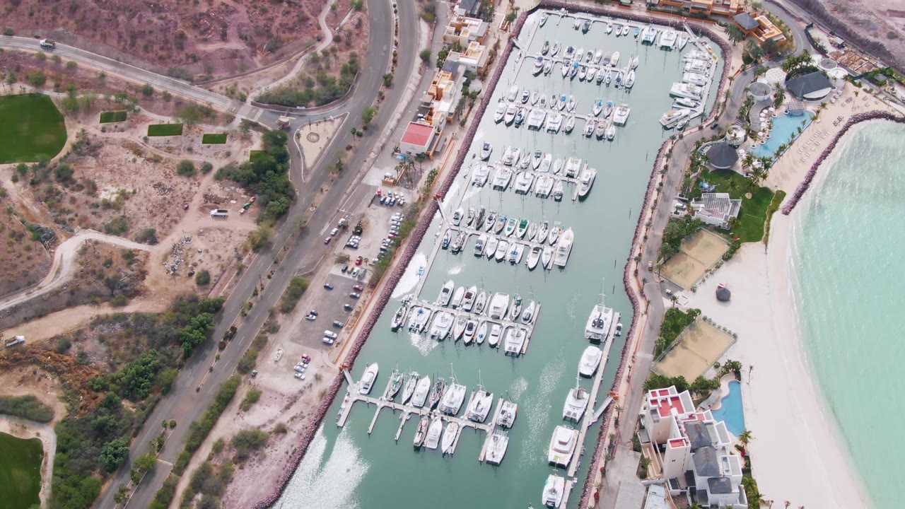 Aerial frame in La Paz, Baja California Sur with the marina centered, road and parking along the left side, beach and pale water to the right, dry hills above, and no visible people.
