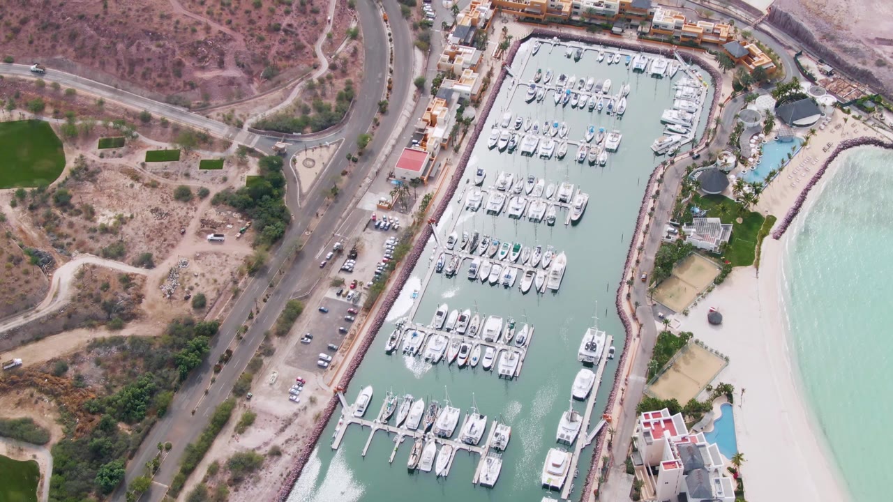 High aerial view in La Paz, Baja California Sur with rows of docked boats, a coastal road with vehicles, a strip of sandy beach, light green water, dry terrain, and no visible people.
