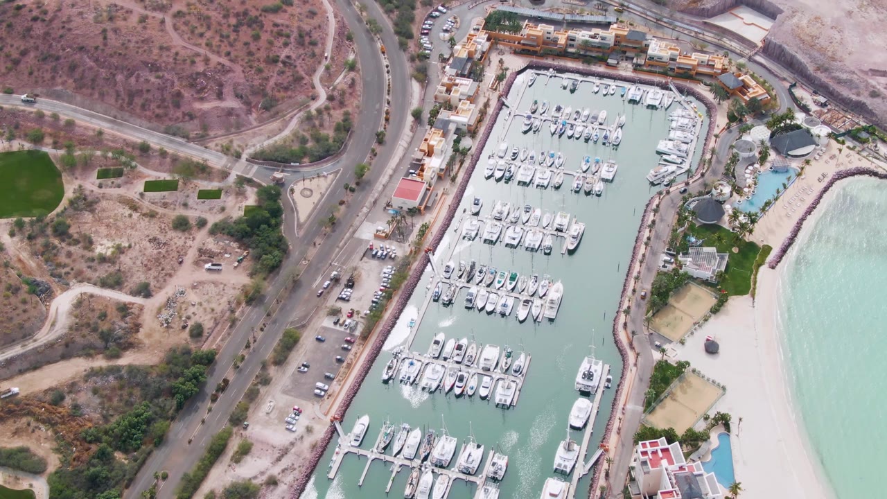 Aerial coastal road curving past marina boats and beach water.