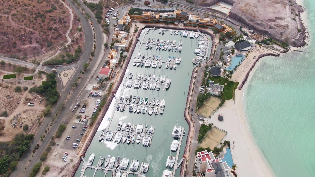 High aerial frame over a La Paz marina with rows of docked boats, sandy beach on the right, road and parked cars on the left, and no visible people.