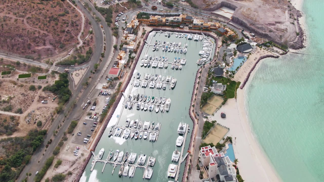 Drone aerial frame in La Paz showing the marina angled below the camera, with docks and boats filling the basin, a sandy beach on the right, and no visible people.