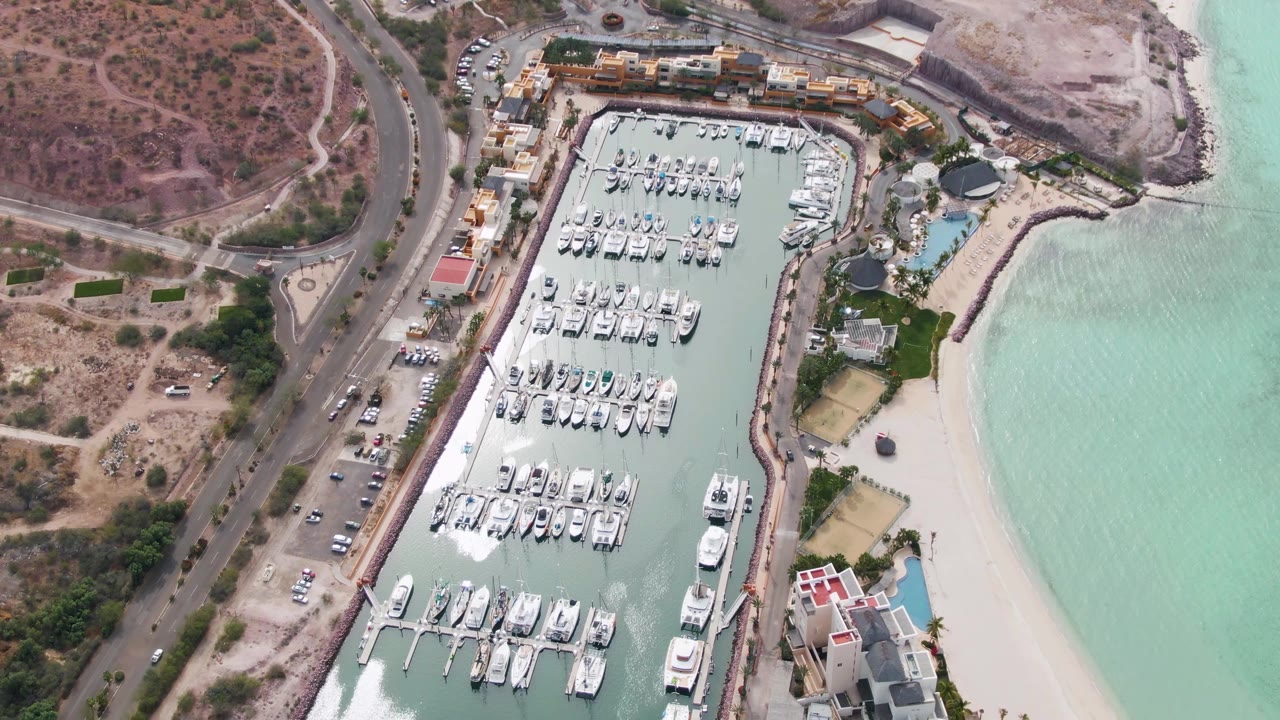 Aerial view of marina slips, white boats, turquoise water, road, parking, and beach.