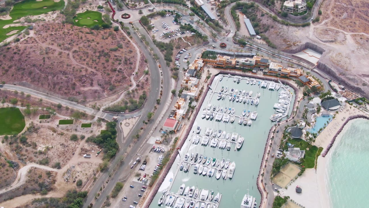Aerial frame over La Paz, Baja California Sur showing a marina filled with docked yachts, a curved coastal road, dry hillside terrain, green patches, beach, and pale water with no visible people.