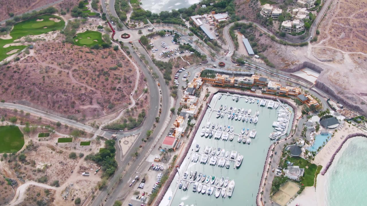 Second aerial frame of La Paz marina with rows of white yachts, vehicles on the curved road, a roundabout near dry terrain, landscaped green areas, and beach water along the lower right, with no visible people.