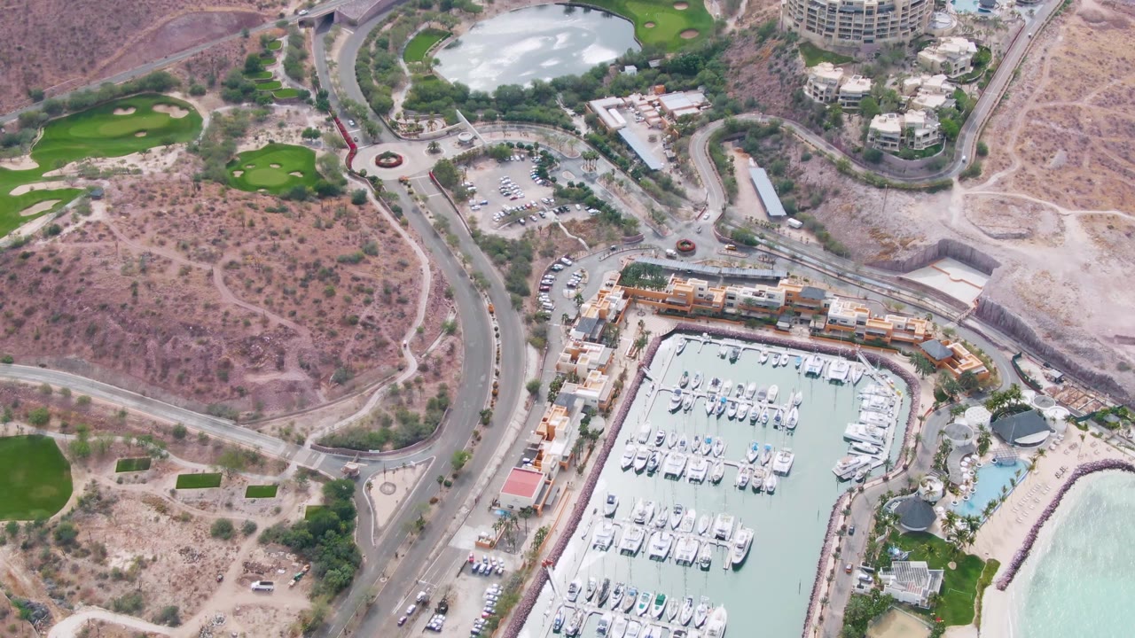 Aerial view in La Paz, Baja California Sur with the marina and docked yachts centered lower in frame, a road and roundabout beside scrubby desert ground, green lawn areas, buildings, and pale shoreline water with no visible people.