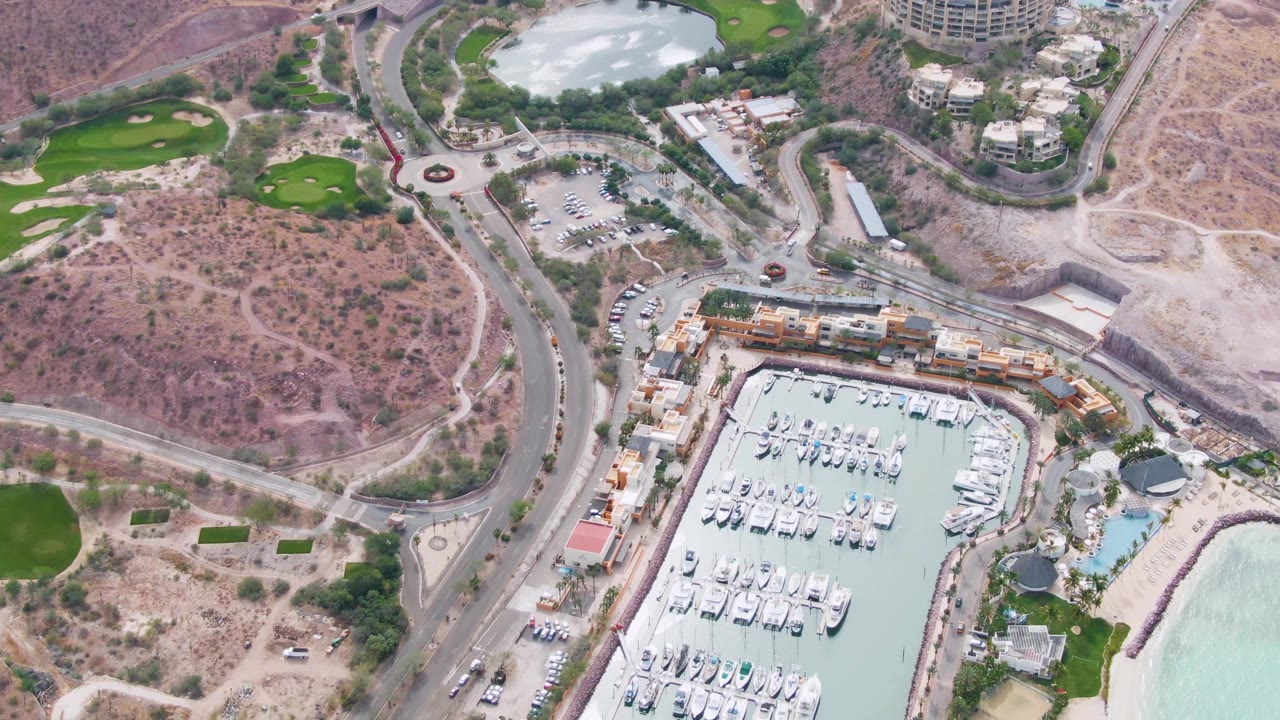 Aerial marina and road view with yachts, roundabout, dry hills, buildings, and beach water.
