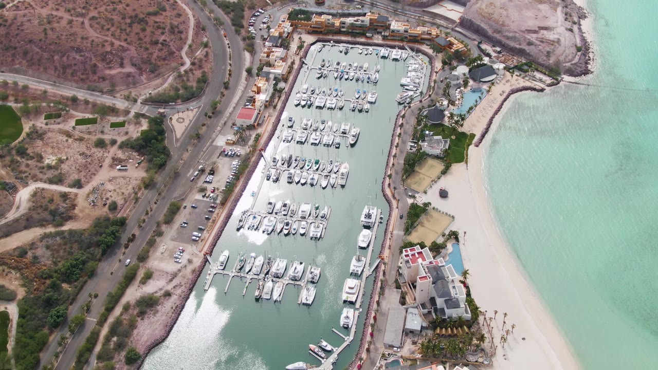 High aerial frame over a La Paz, Baja California Sur marina with rows of docked boats, a sandy beach to the right, a road and dry hillside to the left, and no visible people.