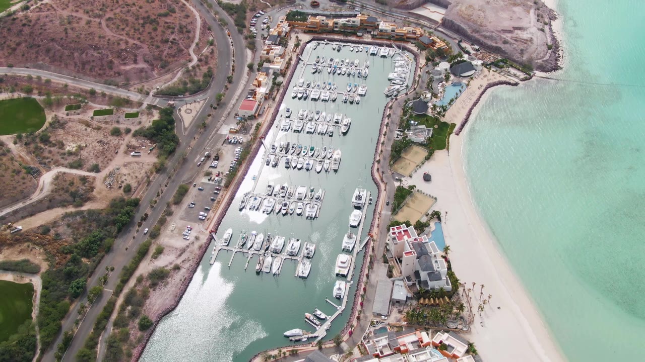 Aerial view in La Paz, Baja California Sur showing the marina shifted slightly lower in frame, with dock rows, beach water, roadside vehicles, coastal buildings, and no visible people.