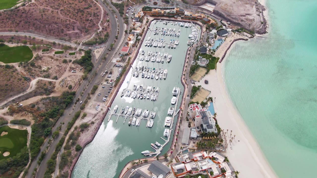 La Paz marina aerial with many white boats docked in rows, a small boat near the lower marina channel, pale beach water on the right, hillside roads on the left, and no visible people.