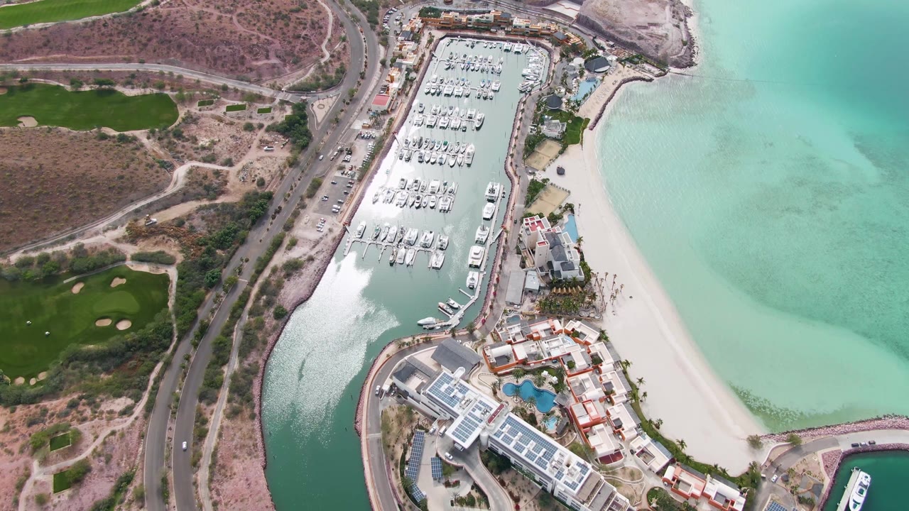 Mid-clip aerial frame over La Paz, Baja California Sur, showing the marina entrance, docked boats, beach, coastal road with small vehicles, golf course greens, and no visible people.