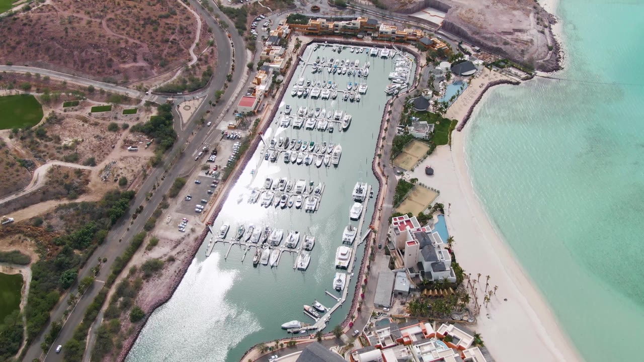 La Paz aerial frame with the boat slips centered, the coastal road and dry terrain along the left edge, sandy beach and pale turquoise shallows on the right, and no visible people.