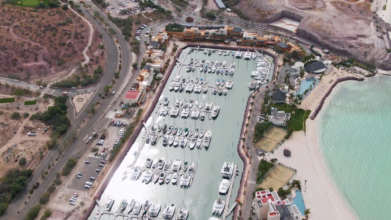 Aerial view of a marina, beach, coastal road, and dry hills.