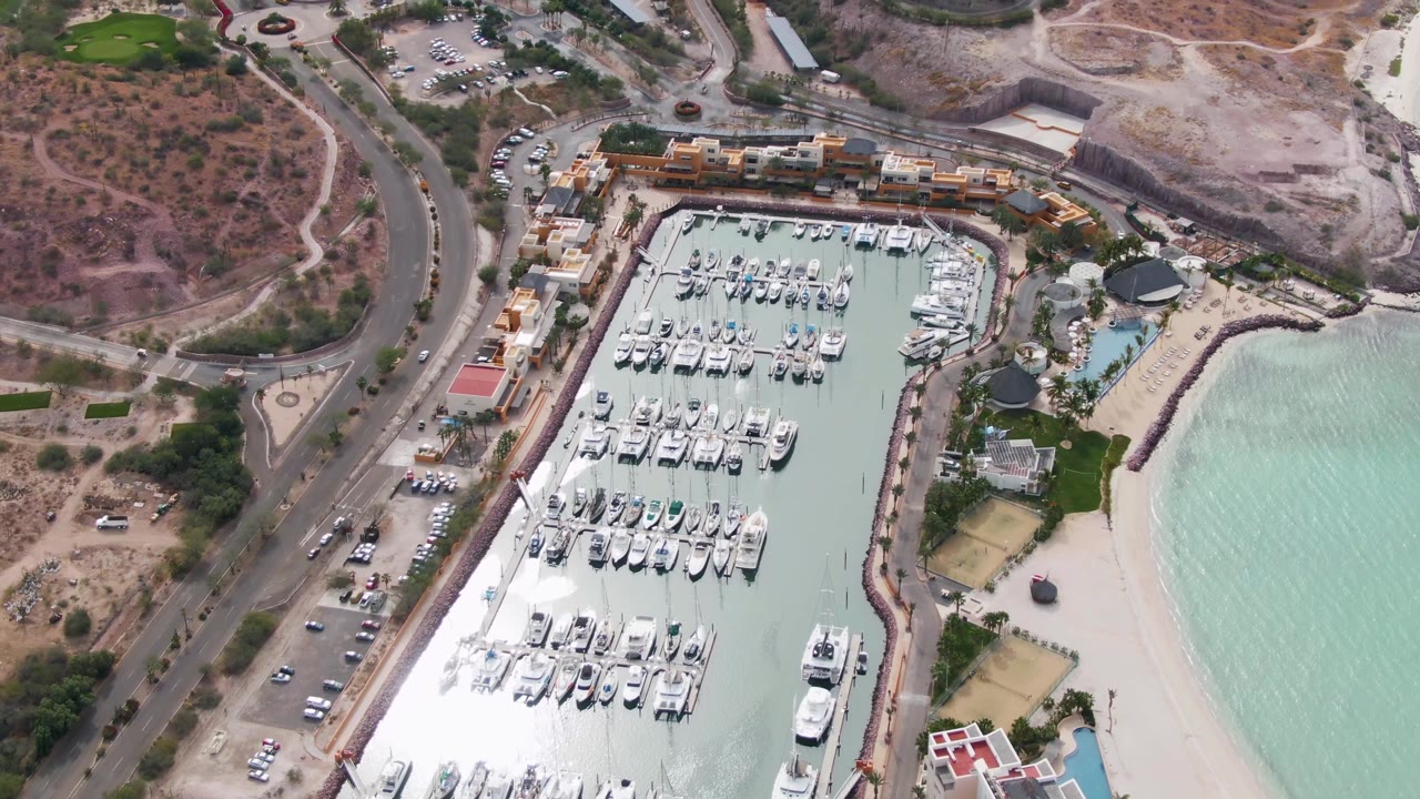 Second aerial frame of the La Paz marina, with dense rows of white boats inside the basin, vehicles along the coastal road, pale beach sand beside clear water, and no visible people.