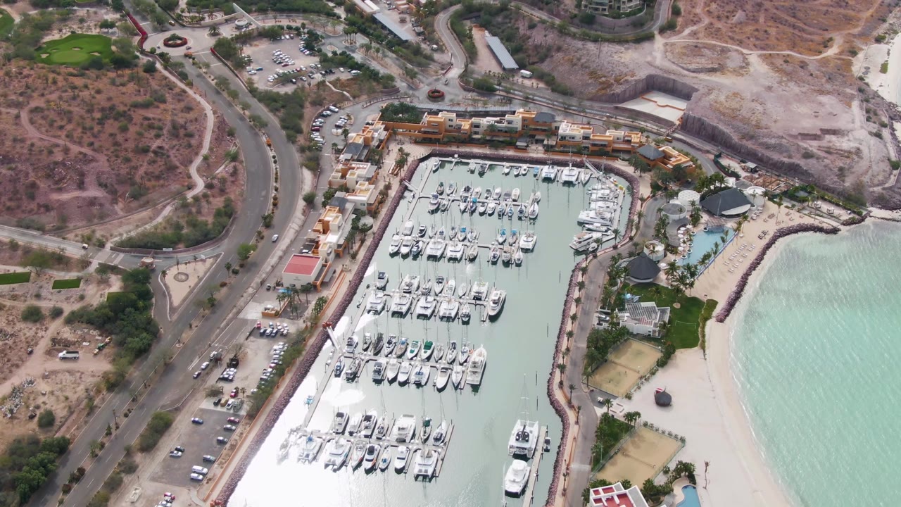 Aerial frame over La Paz showing the marina basin filled with boats, a curved breakwater edge, beach and turquoise water to the right, road and parked vehicles to the left, and no visible people.