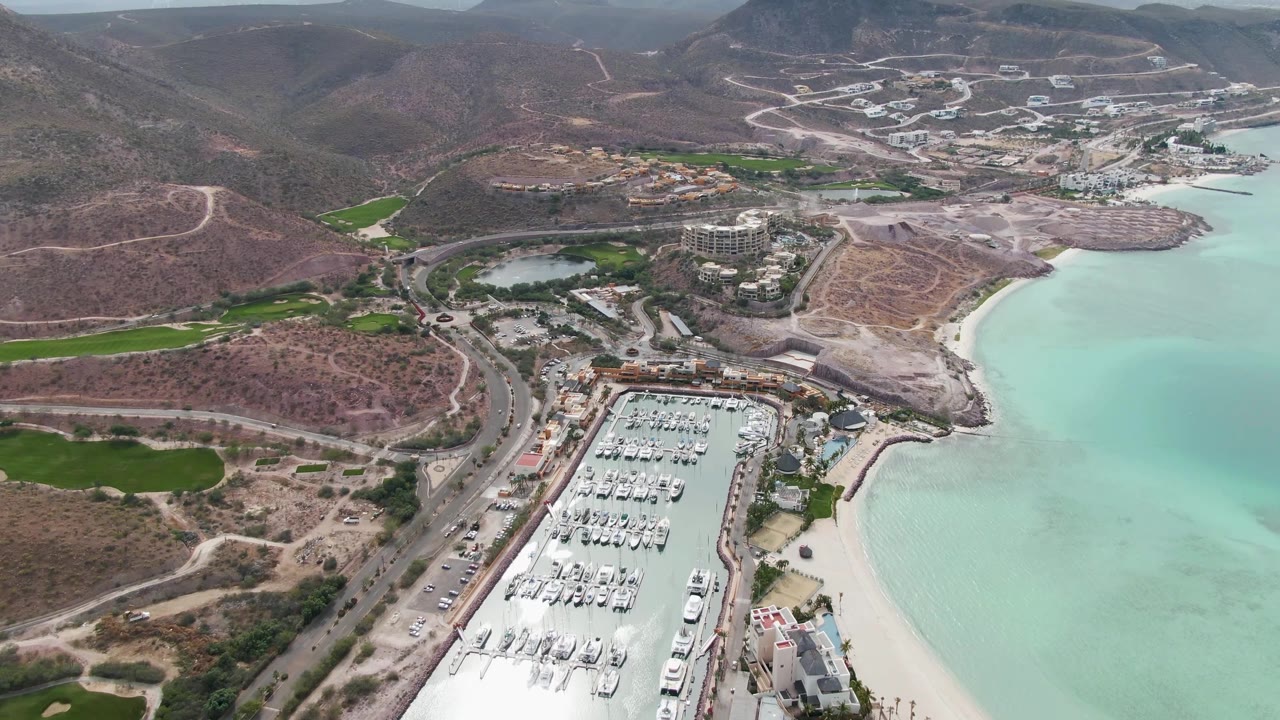 Aerial frame in La Paz, Baja California Sur with the marina centered low in the image, the beach curving along turquoise water, roads crossing dry hills, and no visible people.