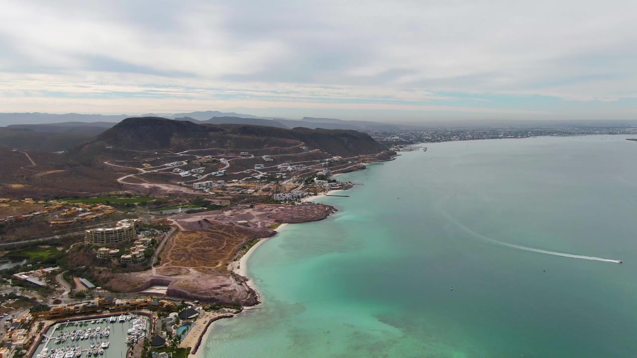 High aerial over La Paz, Baja California Sur with a marina at lower left, curved sandy beach, turquoise shallows, brown hills, coastal buildings, cloudy sky, and no visible people.