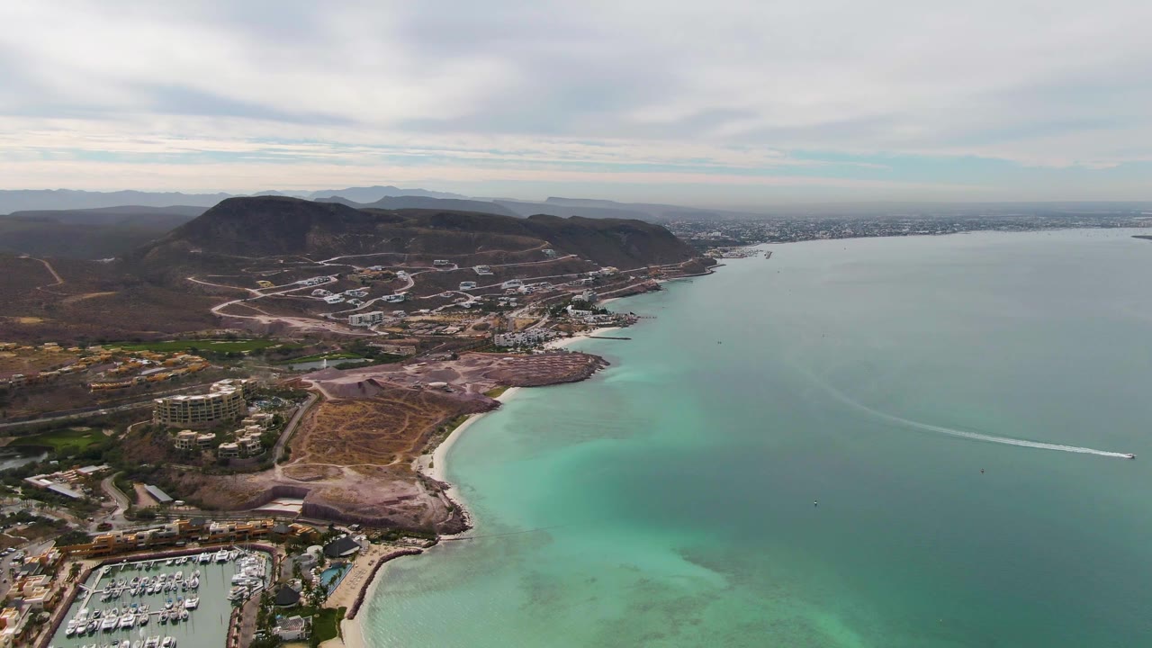 Aerial frame in La Paz showing the marina farther into the lower edge, a pale beach curve, green-blue shallow water, hillside roads, scattered buildings, distant shoreline, and no visible people.