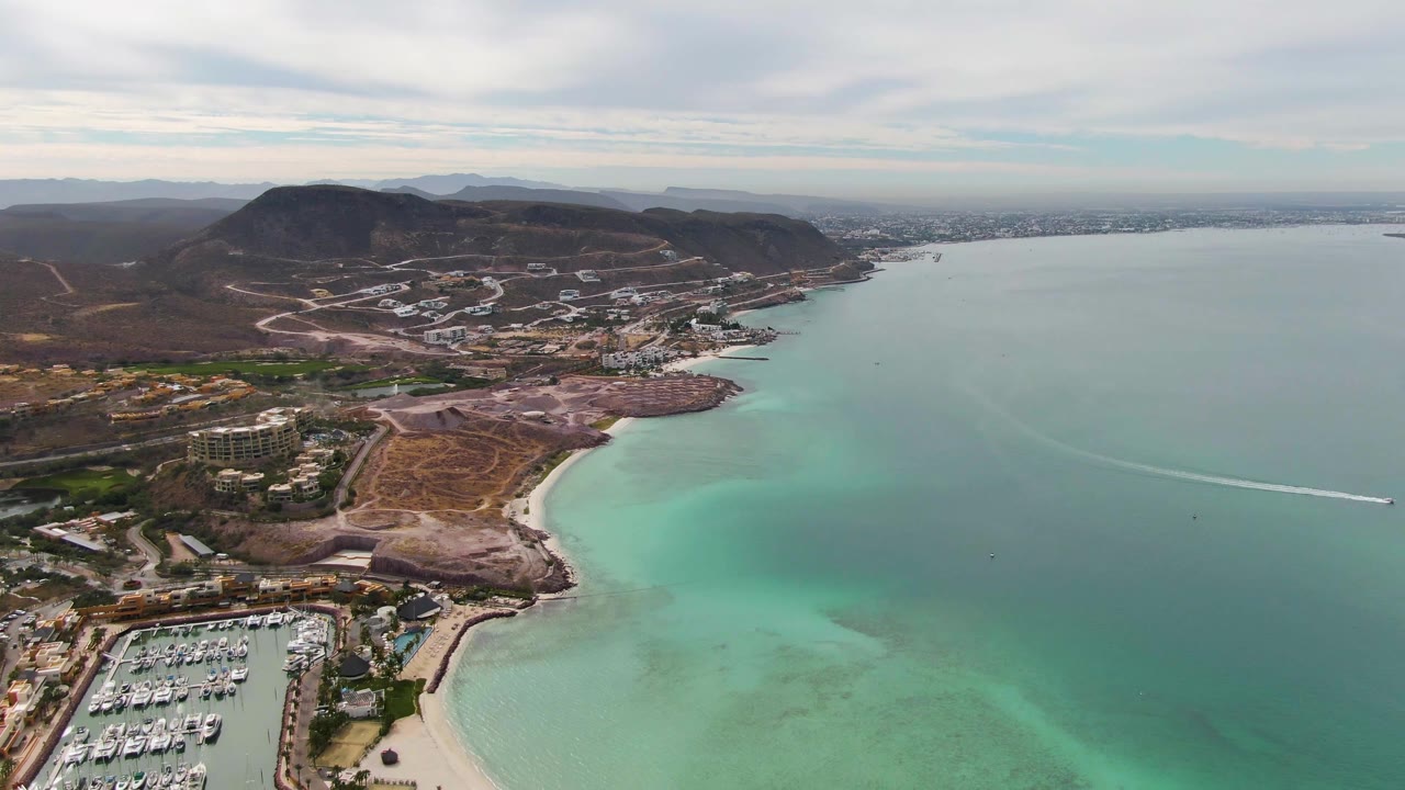 Wide aerial over La Paz with marina slips in the lower left, sandy beach below center, turquoise water across the foreground, brown hills and development inland, a boat wake on the bay, and no visible people.