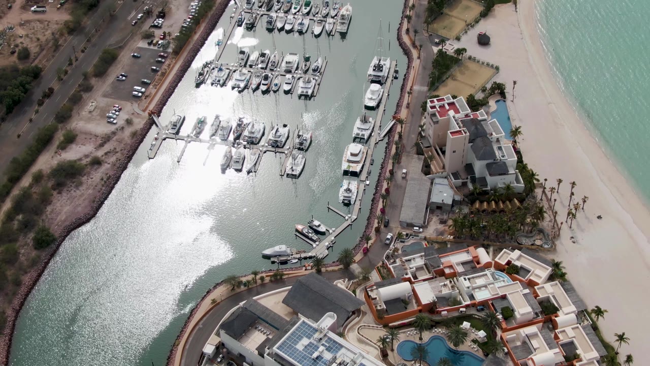 Aerial frame over La Paz, Baja California Sur, showing docked boats, a curved marina edge, pale beach, waterfront buildings, and no visible people.