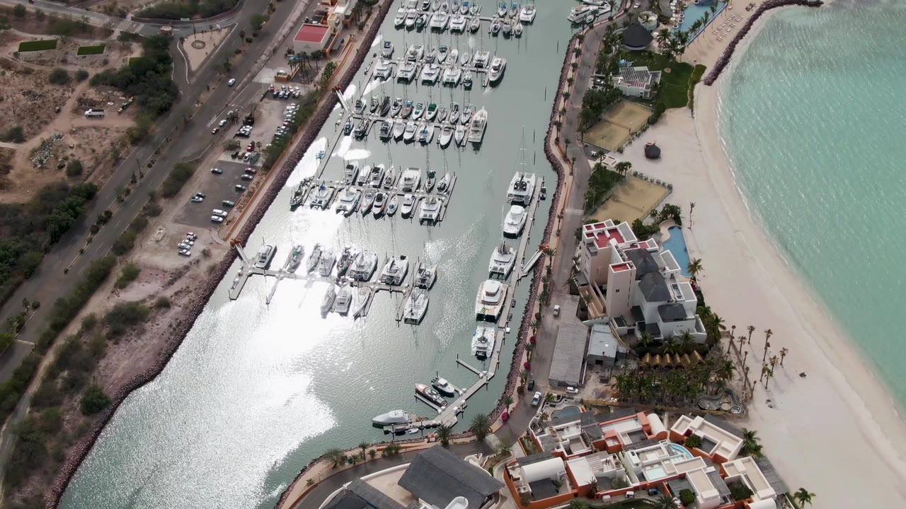 Aerial view of docked boats, bright water reflection, road, beach, and buildings.