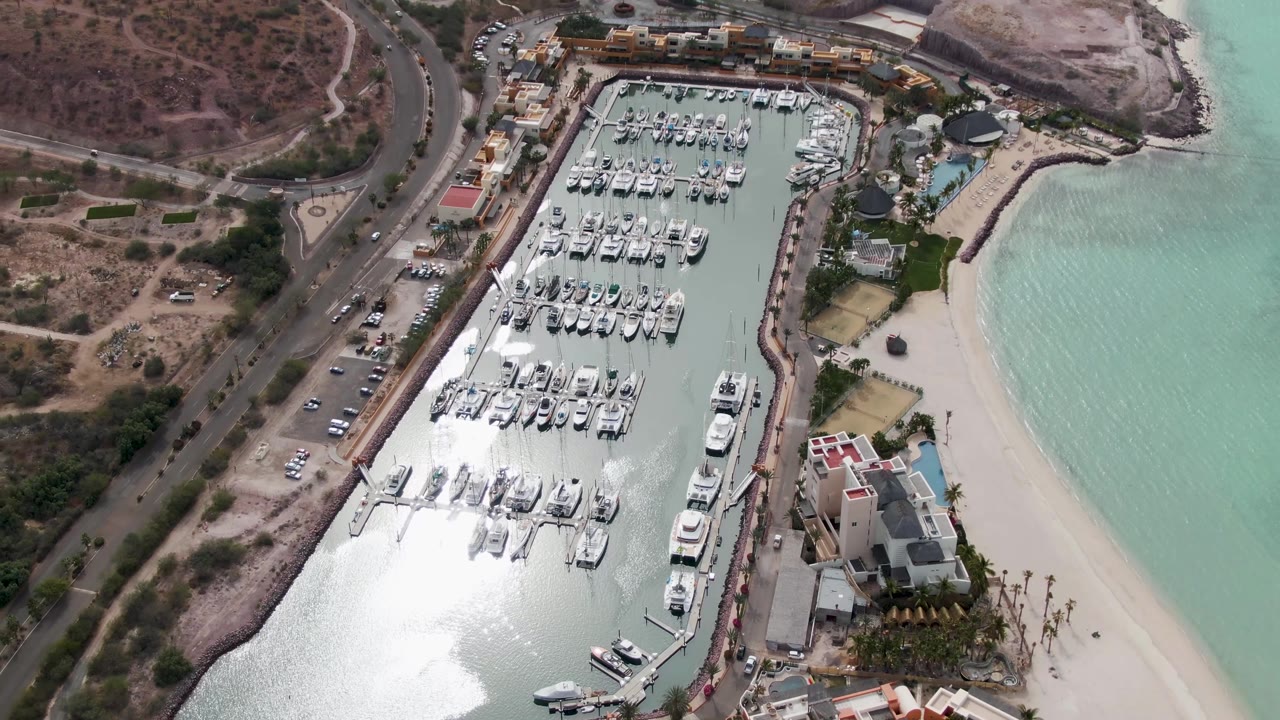 Higher in the La Paz aerial, the marina sits between dry hillside roads and turquoise shoreline, with docked boats, beach sand, waterfront buildings, and no visible people.