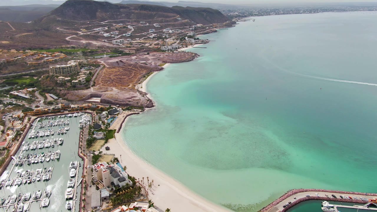 Opening aerial frame over La Paz, Baja California Sur, showing a packed marina at lower left, pale beach, turquoise bay water, hillside roads, and no visible people.
