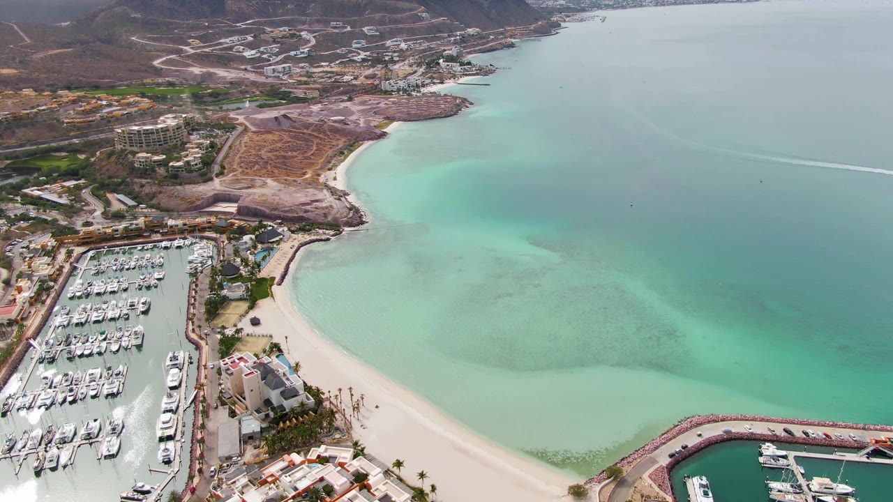 Aerial frame in La Paz with the marina shifted lower left, a curved white sand beach through the center, clear shallow water to the right, and no visible people.