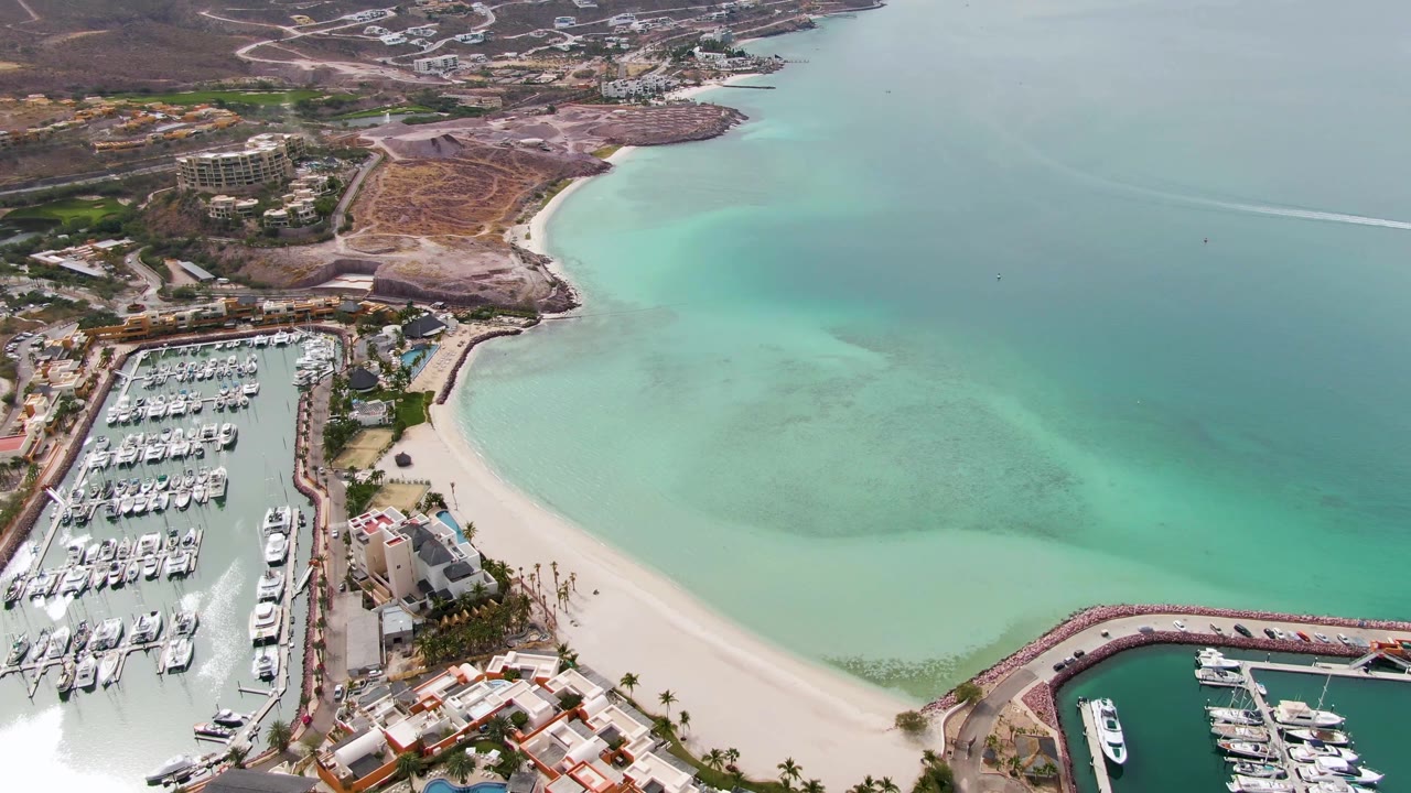 La Paz aerial frame showing more open turquoise water at right, marina docks at left, a beach arc below, shoreline buildings, and no visible people.