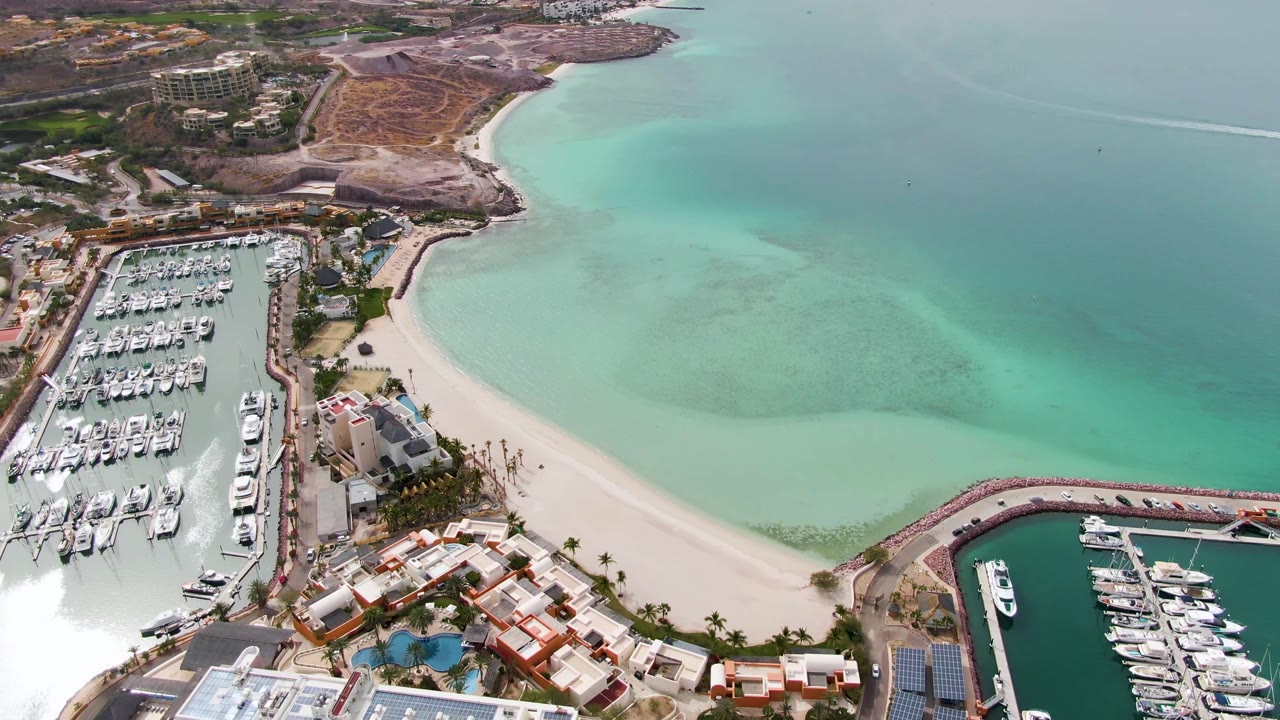 Aerial view of marina basins, beach, palm-lined buildings, and calm turquoise water.
