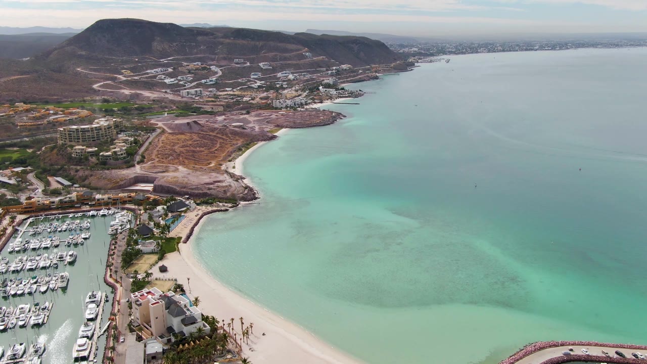 Aerial frame over La Paz, Baja California Sur showing a marina at lower left, pale beach, turquoise bay water, hillside roads, scattered buildings, brown hills, and no visible people.