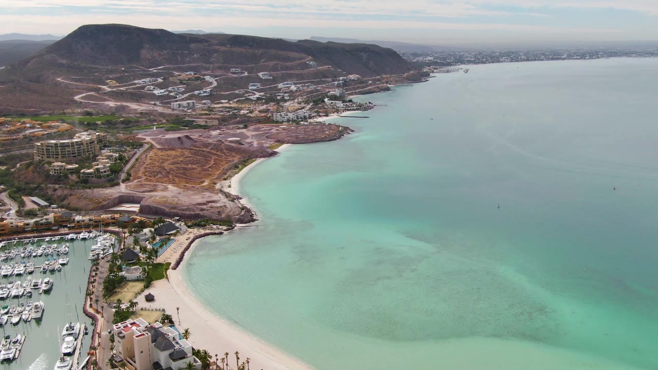 Aerial frame over La Paz, Baja California Sur with the marina still visible on the left edge, a curved sandy beach below, shallow turquoise water across the bay, hillside roads, and no visible people.