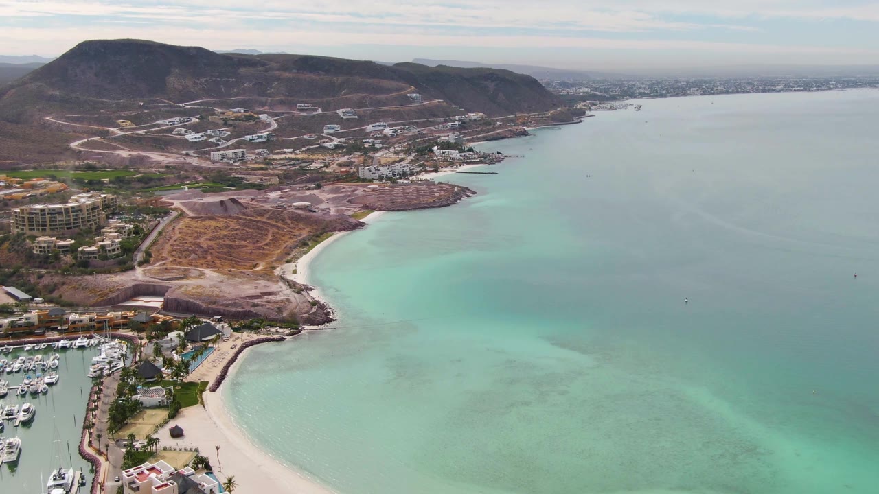 Aerial frame over La Paz, Baja California Sur showing the beach and marina pushed toward the left edge, broad turquoise-green water in the foreground, hillside development beyond the shore, and no visible people.