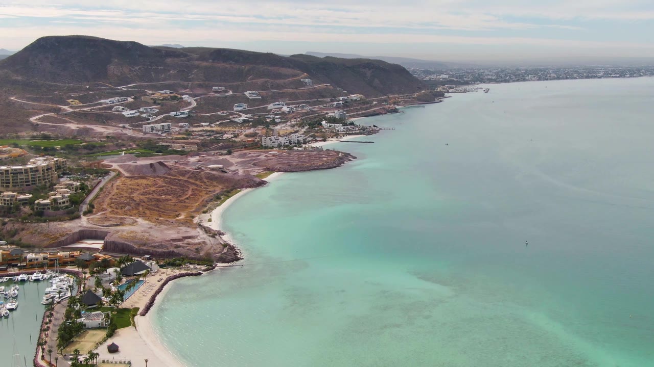 Aerial view of turquoise bay, curved beach, marina edge, hillside roads, and brown hills.
