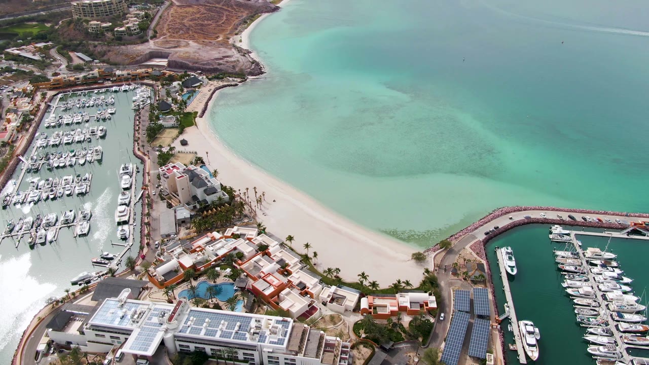 Aerial frame over La Paz, Baja California Sur showing two marina basins, rows of docked boats, a curved sandy beach, turquoise water, nearby buildings, and no visible people.