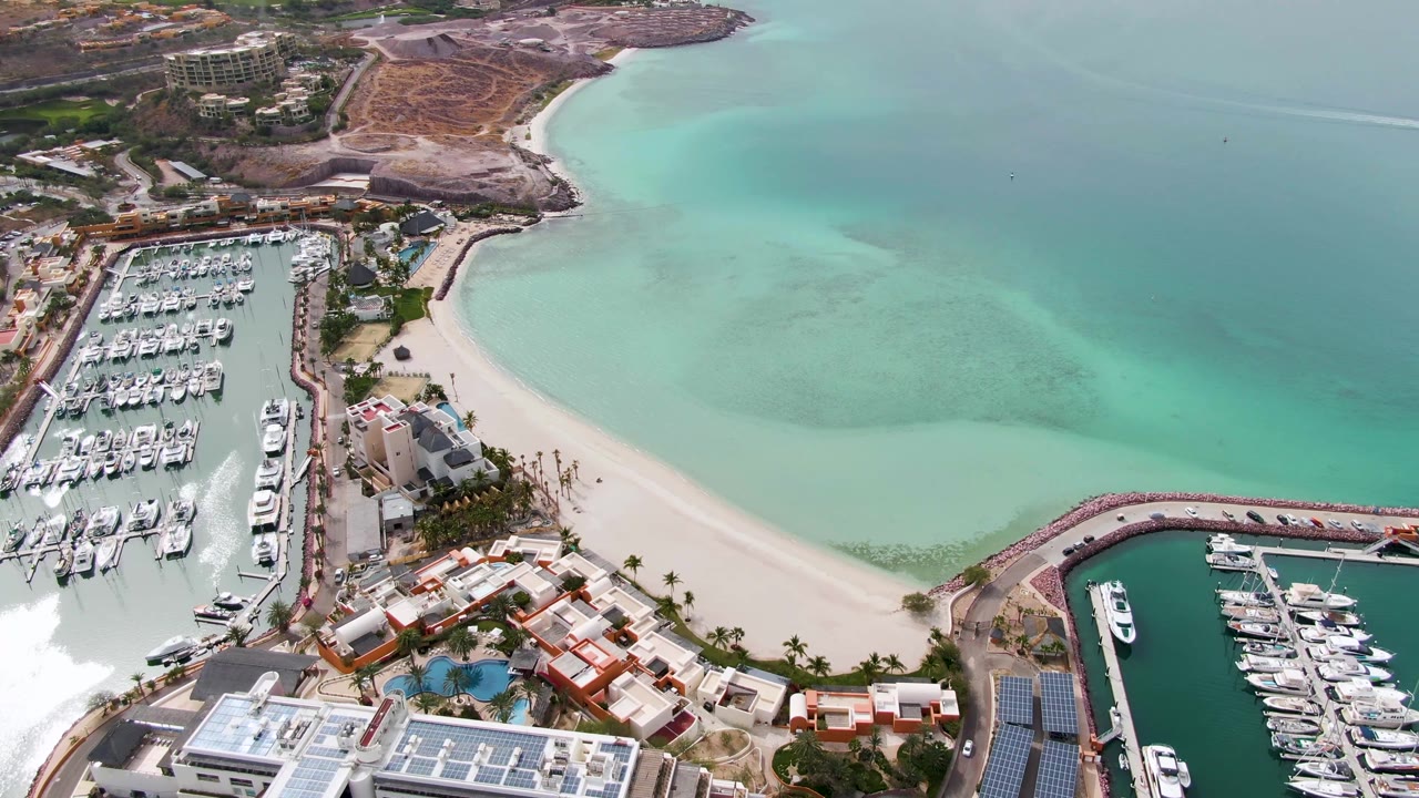 One second into the La Paz aerial, docked yachts fill the left marina while a pale sandy beach curves around turquoise water, with no visible people.