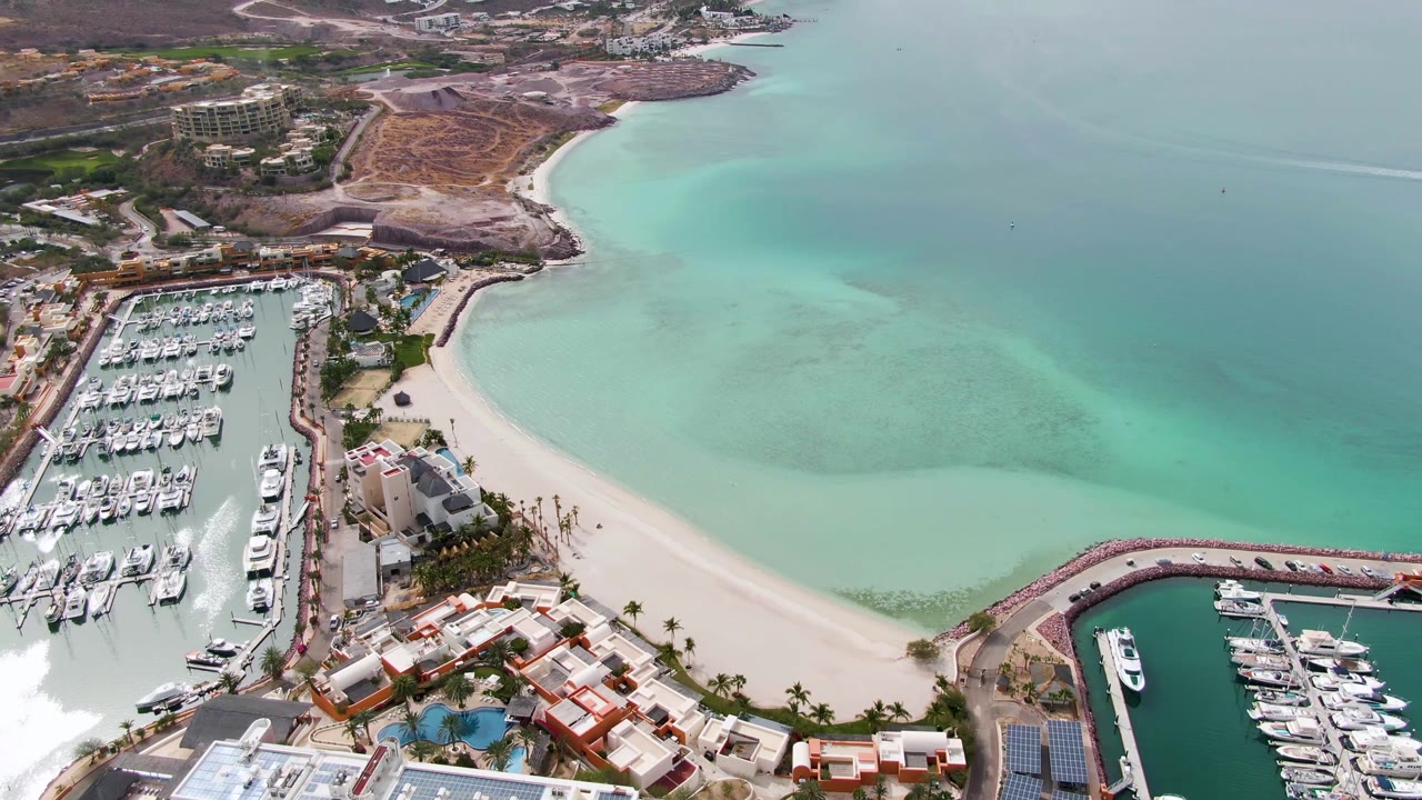 La Paz aerial view shows marina slips at left and lower right, a light sand beach between them, turquoise bay water, roads, hillside development, and no visible people.
