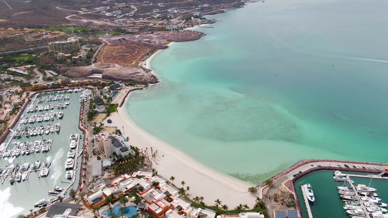 Aerial coastal view of a marina, sandy beach, turquoise water, and dry hills.