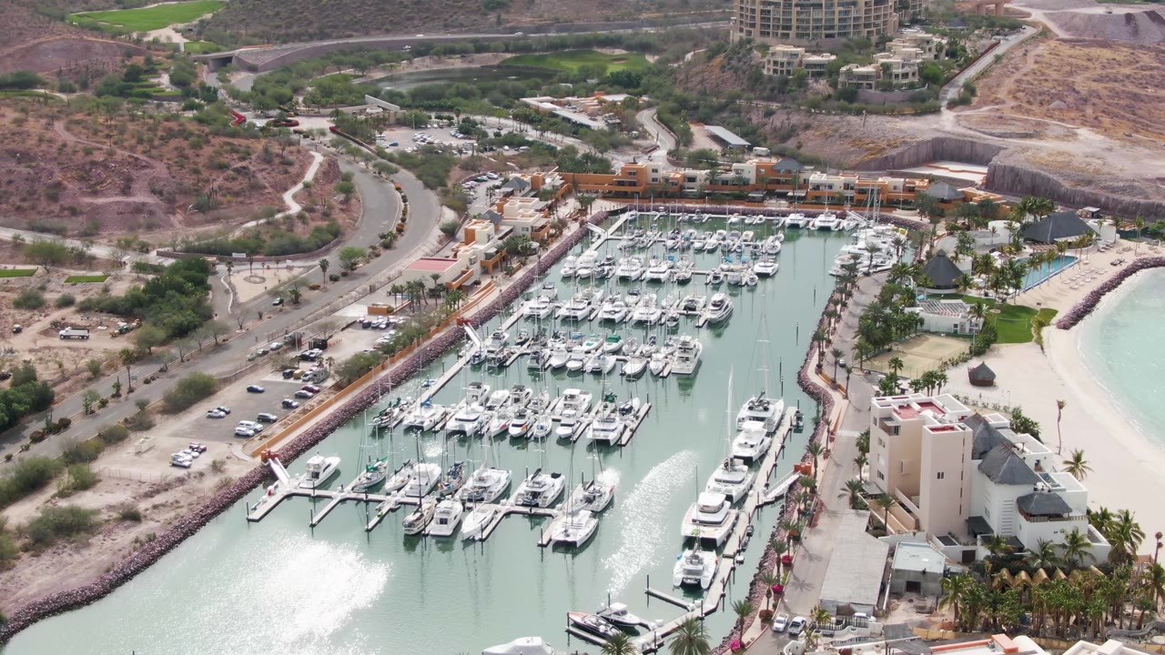 High aerial view over a La Paz, Baja California Sur marina with rows of docked white boats, pale green water, sandy beach, waterfront buildings, a curved road, dry hills, and no visible people.