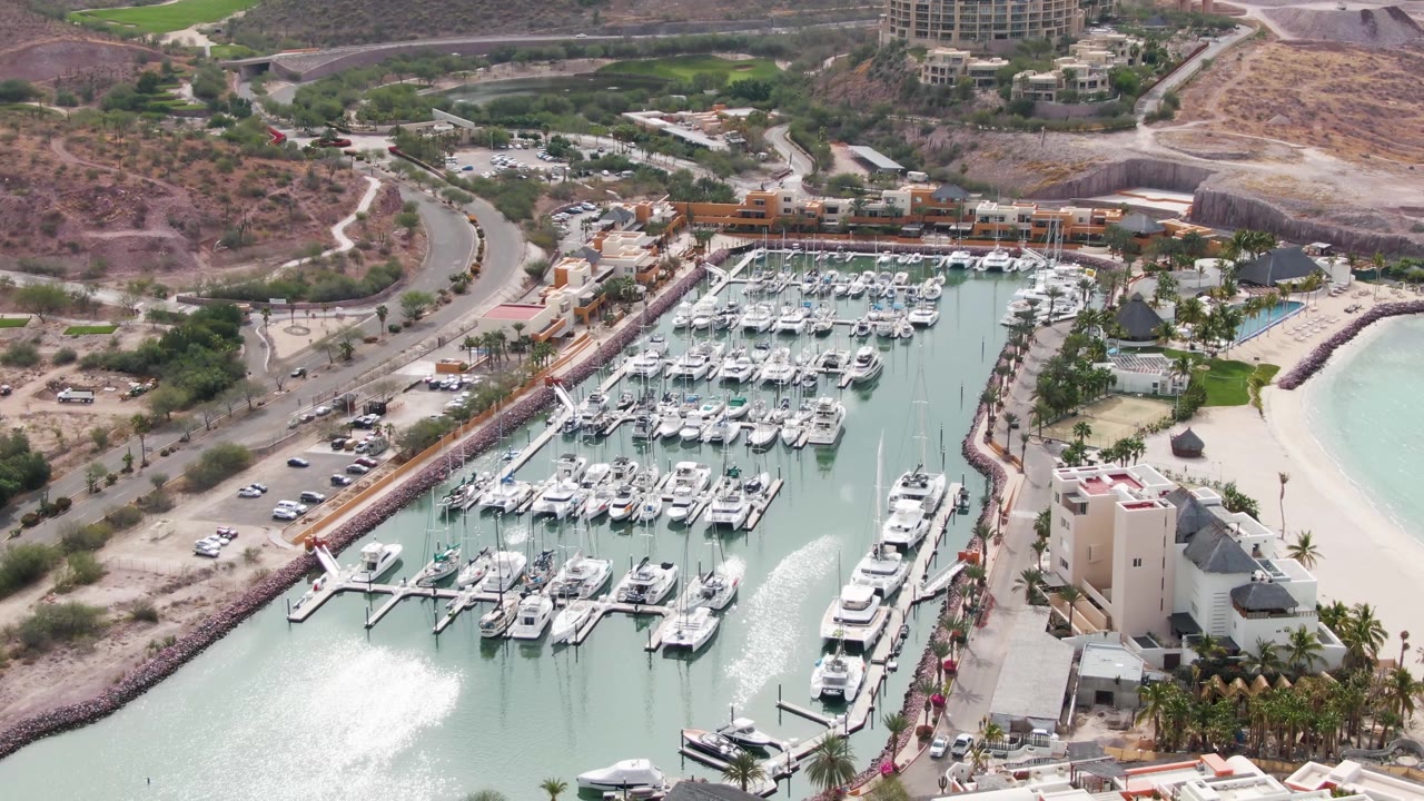 A slightly later aerial frame shows the La Paz marina centered with dense rows of boats, a bright beach edge at right, waterfront structures below, road traffic at left, dry hills beyond, and no visible people.