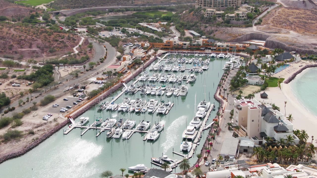 Aerial view of the La Paz marina with the boat slips filling the center, a pale sandy beach and light water to the right, a road and parked vehicles to the left, hillside terrain above, and no visible people.
