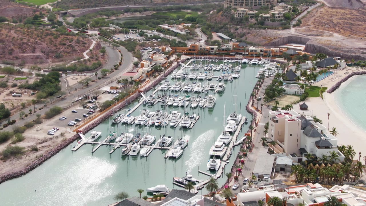 Aerial view of marina docks, yachts, beach, buildings, vehicles, and hills.
