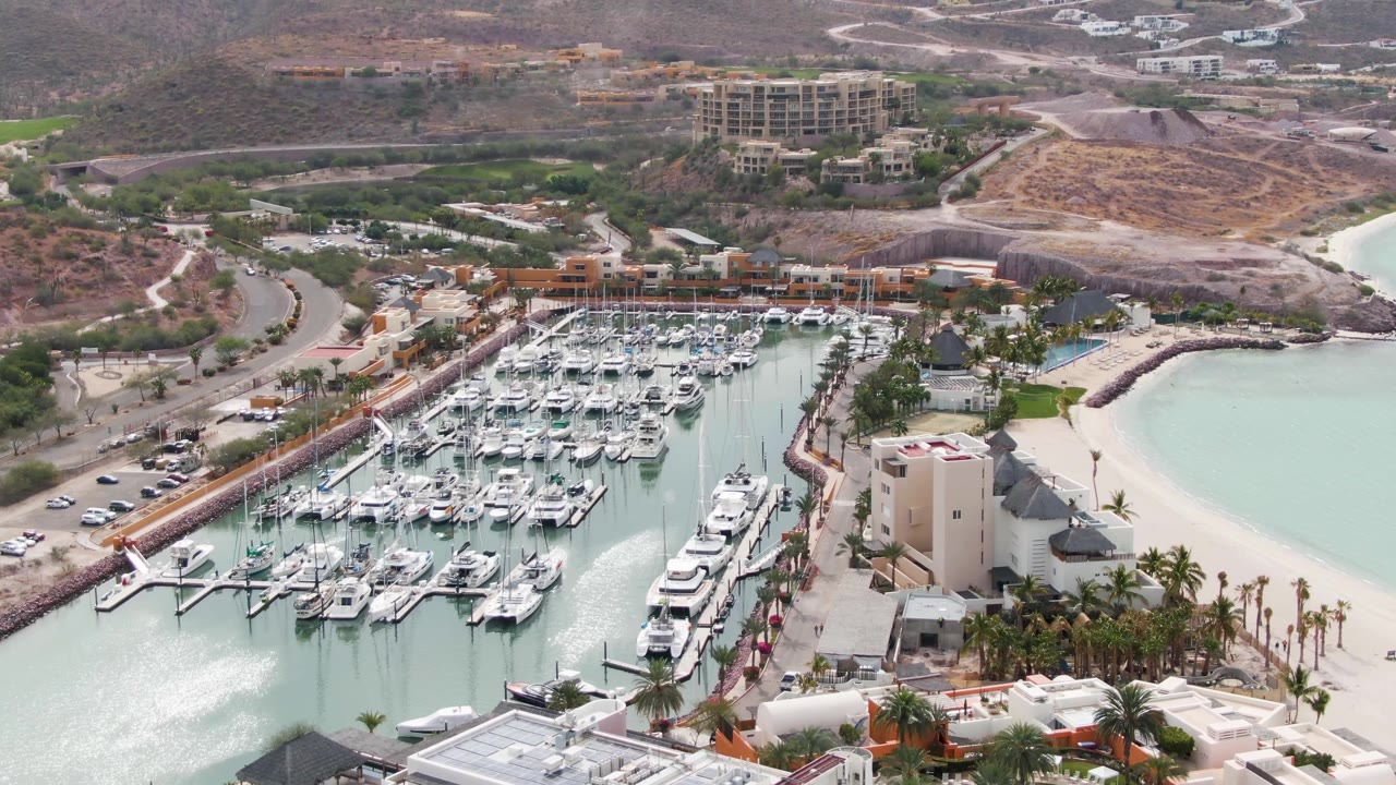 Aerial frame over La Paz, Baja California Sur showing packed marina docks, a sandy beach, palm trees, nearby roads, hillside buildings, pale turquoise water, and no visible people.