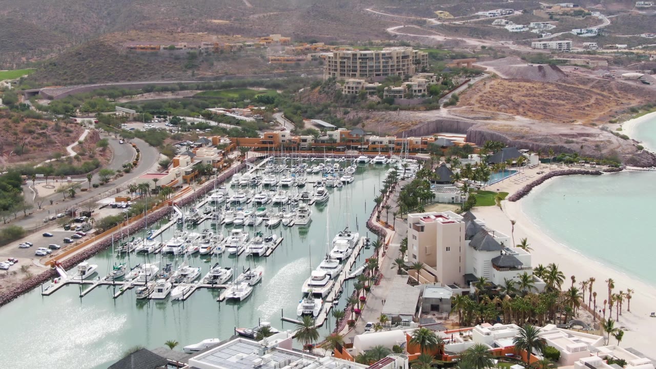 La Paz aerial frame with the marina centered, white boats clustered on docks, a curved sandy beach to the right, palms along the shore, roads and hills behind, and no visible people.