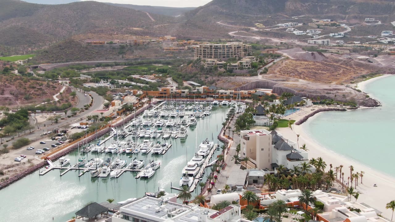 A high aerial frame in La Paz shows rows of marina slips and yachts, a palm-lined shore, the pale beach edge, resort-like buildings, roads, brown hills, and no visible people.