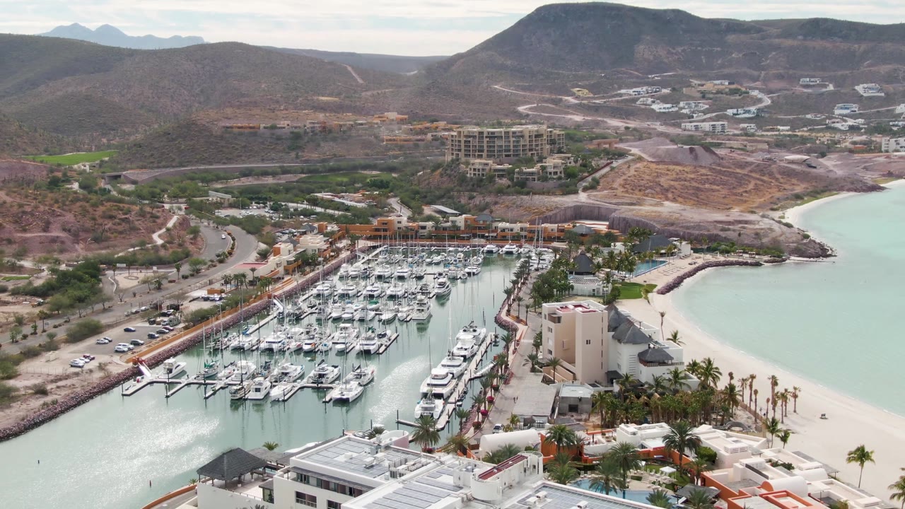 Aerial frame with marina boats, shoreline buildings, pale water, roads, and mountains.