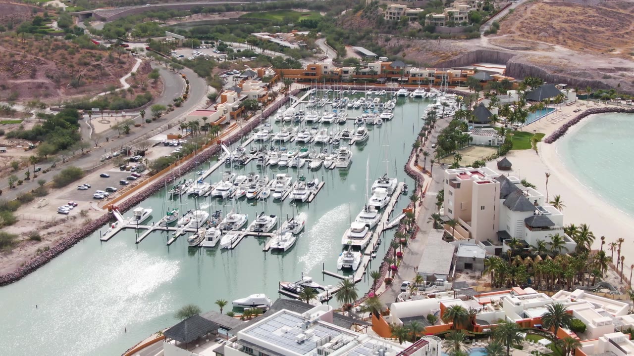 Opening aerial frame over a La Paz, Baja California Sur marina with rows of white boats, pale green water, a curved beach, road traffic, hillside buildings, and no visible people.